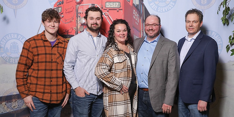 Cole, Jaden, Julie, Jesse and Ben Chalich of Northern Plains Railroad posing for a photo in front of a mural of a bright red locomotive engine.