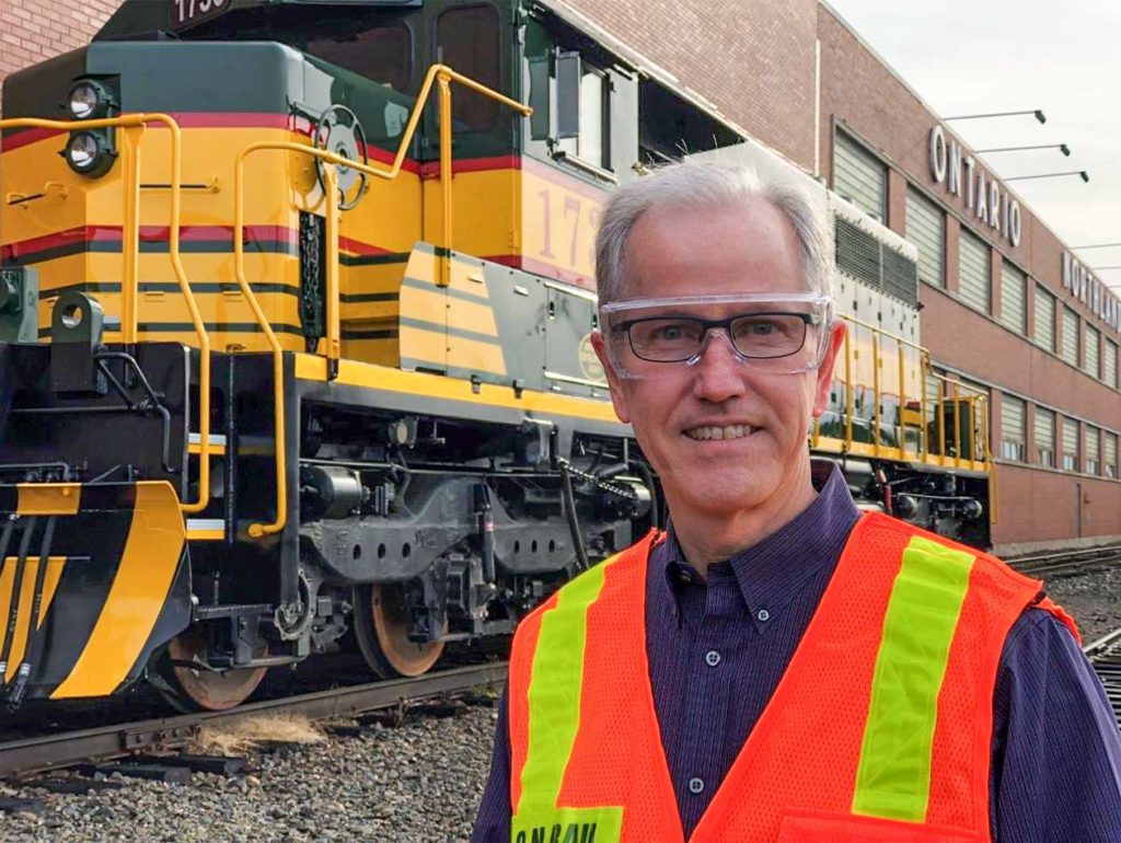 Northern Plains Railroad Services engineer Grant Bailey smiling for a photo with a locomotive engine behind him.