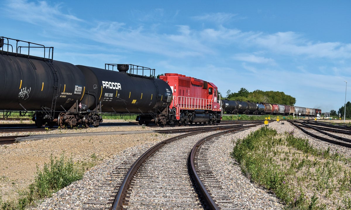 A red locomotive with two black tanker cars behind it, with a railroad track in the foreground.