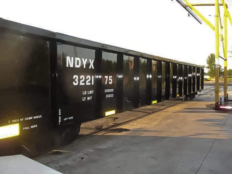 A shiny black open-top hopper car, freshly refurbished, sits on a track in the sunshine.